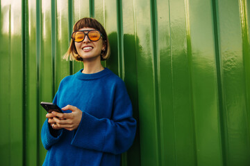 Happy young caucasian woman smiles with teeth looking at camera, holding phone standing against green wall. Brown-haired with bob haircut wears blue sweater in autumn weather. City life concept