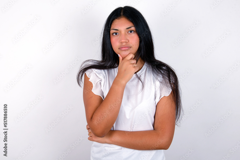 Thoughtful smiling young brunette woman wearing white T-shirt standing against white background keeps hand under chin, looks directly at camera, listens something with interest. Youth concept. Thoughtful smiling young brunette woman wearing white T-shirt standing against white background keeps hand under chin, looks directly at camera, listens something with interest. Youth concept.