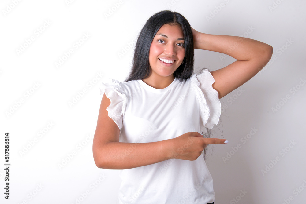 young brunette woman wearing white T-shirt standing against white background feeling positive has amazed expression, indicates something. One hand on head and pointing with other hand. young brunette woman wearing white T-shirt standing against white background feeling positive has amazed expression, indicates something. One hand on head and pointing with other hand.