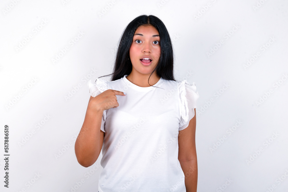 young brunette woman wearing white T-shirt standing against white background being in stupor shocked, has astonished expression pointing at oneself with finger saying: Who me? young brunette woman wearing white T-shirt standing against white background being in stupor shocked, has astonished expression pointing at oneself with finger saying: Who me?