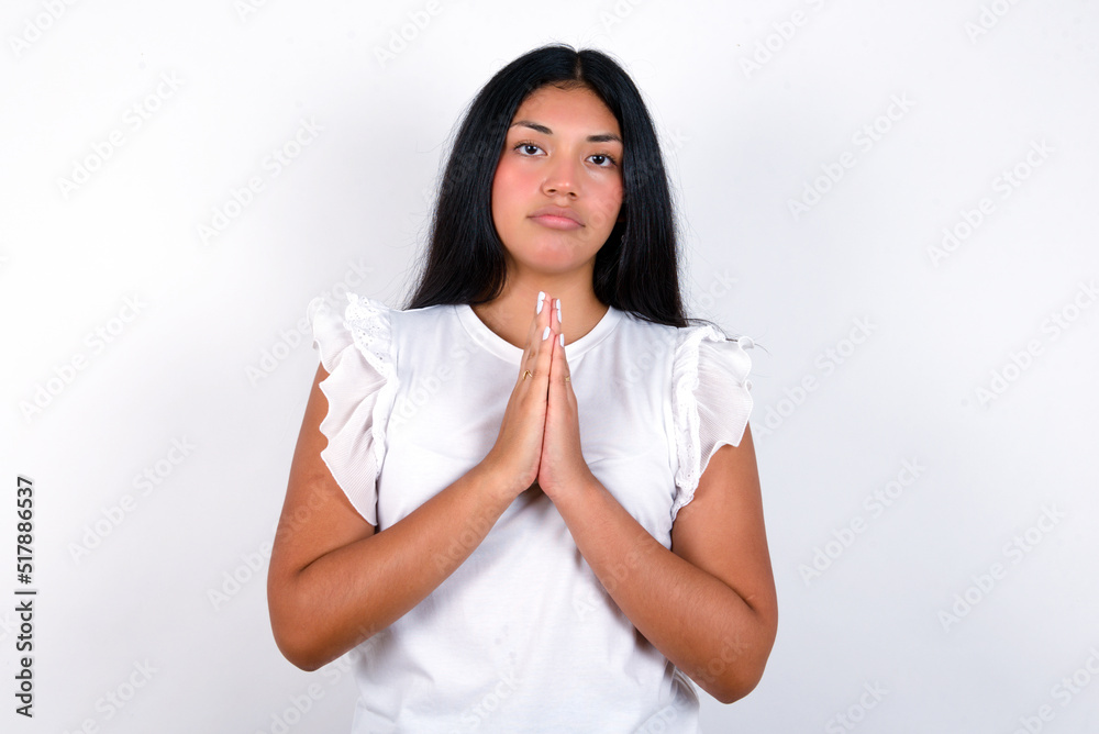 young brunette woman wearing white T-shirt standing against white background keeps palms pressed together in front of her having regretful look, asking for forgiveness. Forgive me please. young brunette woman wearing white T-shirt standing against white background keeps palms pressed together in front of her having regretful look, asking for forgiveness. Forgive me please.