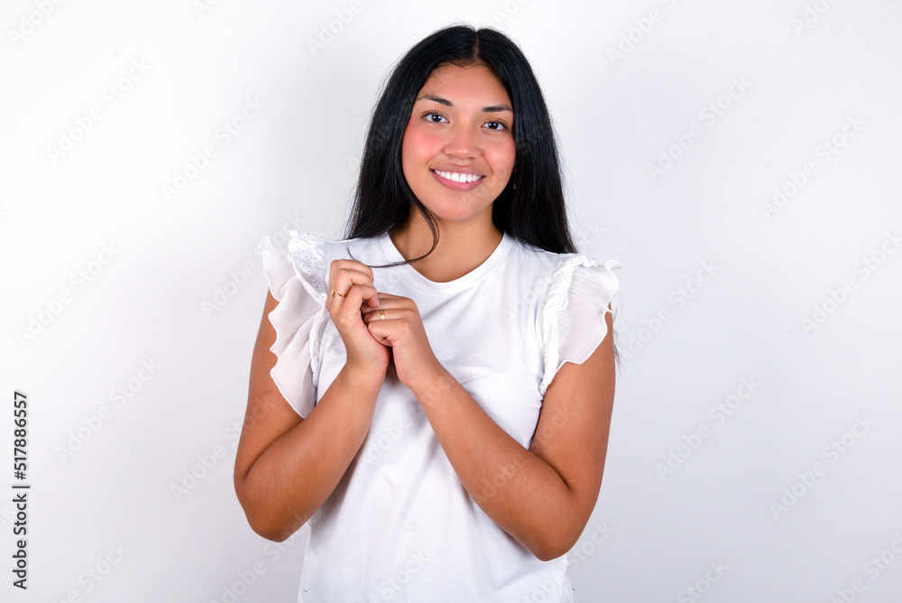 Dreamy charming young brunette woman wearing white T-shirt standing against white background with pleasant expression, keeps hands crossed near face, excited about something pleasant. Dreamy charming young brunette woman wearing white T-shirt standing against white background with pleasant expression, keeps hands crossed near face, excited about something pleasant.
