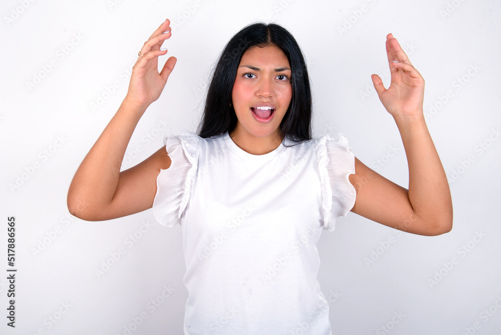 young brunette woman wearing white T-shirt standing against white background looks with excitement at camera, keeps hands raised over head, notices something unexpected, reacts on sudden news.