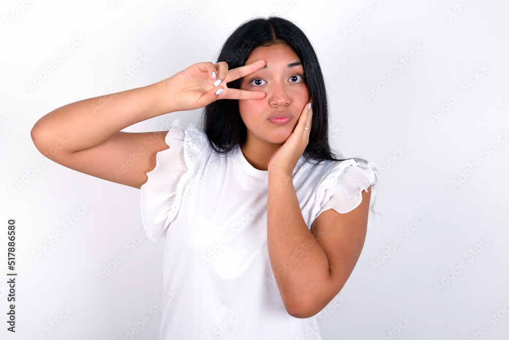 young brunette woman wearing white T-shirt standing against white background making v-sign near eyes. Leisure lifestyle people person celebrate flirt coquettish concept. young brunette woman wearing white T-shirt standing against white background making v-sign near eyes. Leisure lifestyle people person celebrate flirt coquettish concept.