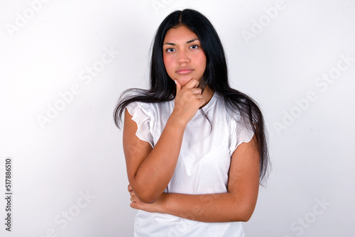 Thoughtful smiling young brunette woman wearing white T-shirt standing against white background keeps hand under chin, looks directly at camera, listens something with interest. Youth concept. © Jihan