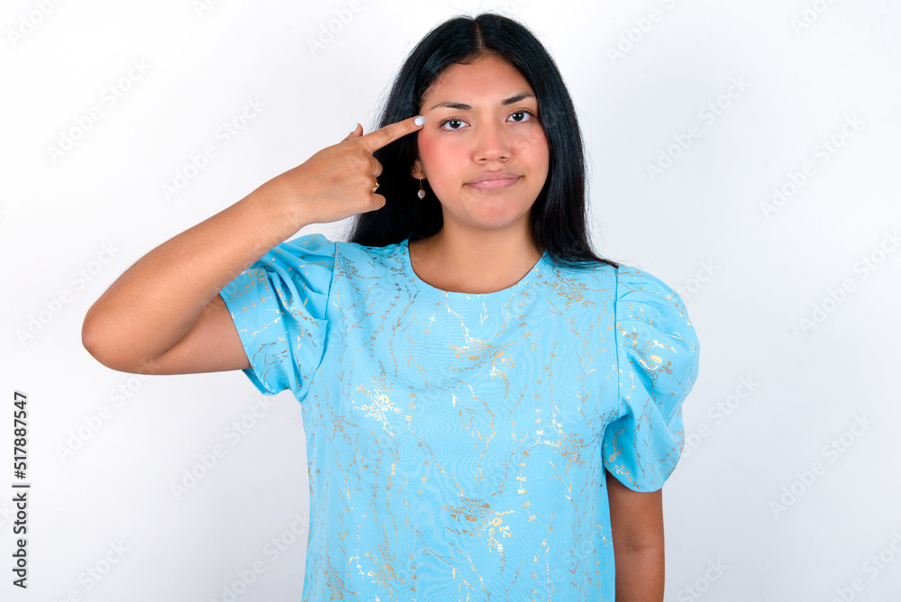 Unhappy young brunette woman wearing blue T-shirt standing against white background curves lips and makes suicide gesture, shoots in temple with hand, tries kill himself. Unhappy young brunette woman wearing blue T-shirt standing against white background curves lips and makes suicide gesture, shoots in temple with hand, tries kill himself.