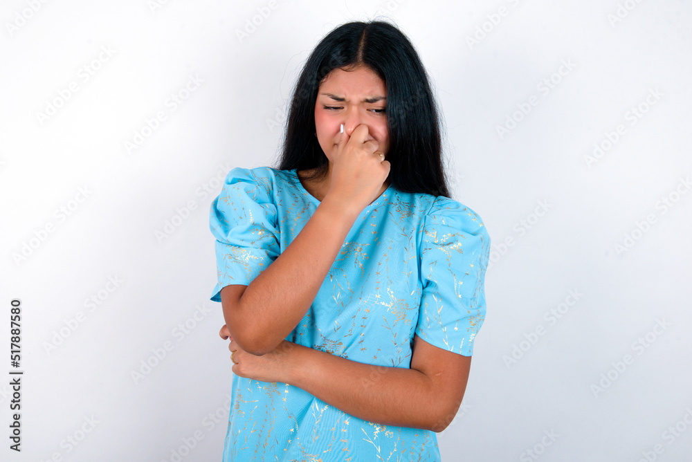 young brunette woman wearing blue T-shirt standing against white background, holding his nose because of a bad smell. young brunette woman wearing blue T-shirt standing against white background, holding his nose because of a bad smell.