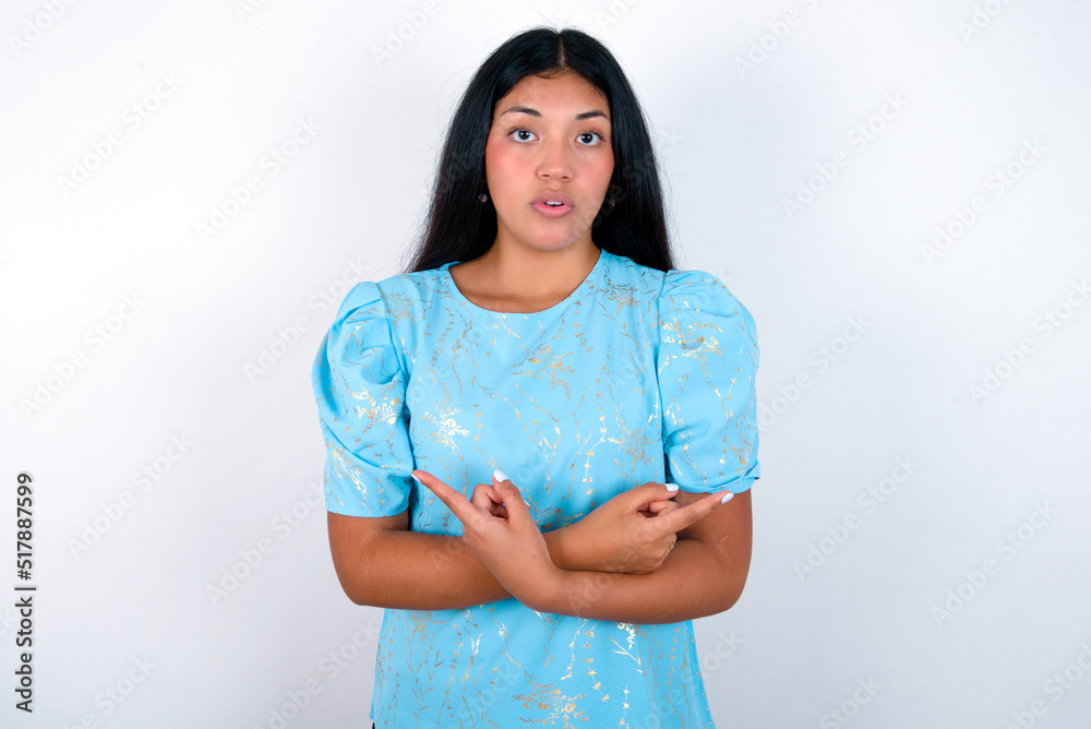 Confused young brunette woman wearing blue T-shirt standing against white background chooses between two ways, points at both sides with crossed hands, feels doubt. Need your advice. Confused young brunette woman wearing blue T-shirt standing against white background chooses between two ways, points at both sides with crossed hands, feels doubt. Need your advice.