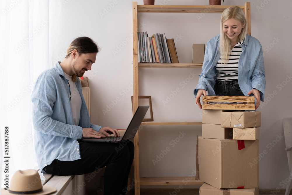Young couple moving in new home. Happy homeowners sitting in a apartment using a laptop. A woman is unpacking things. Searching home decorating ideas on a notebook Young couple moving in new home. Happy homeowners sitting in a apartment using a laptop. A woman is unpacking things. Searching home decorating ideas on a notebook