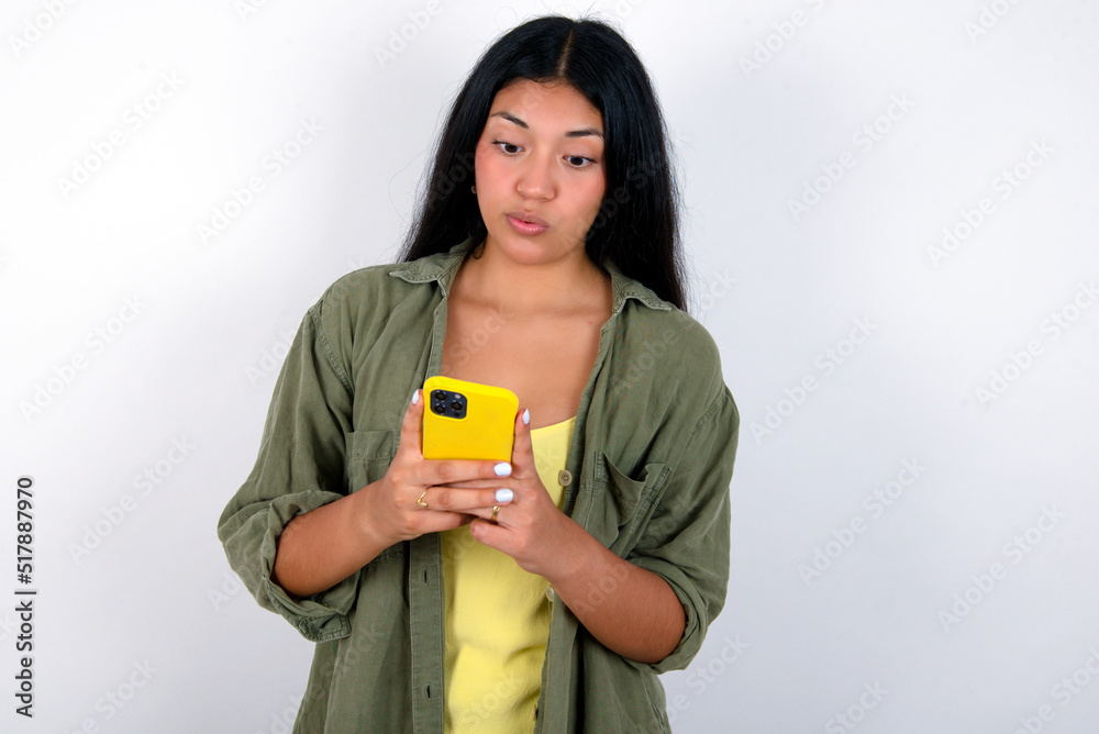 young brunette woman wearing green overshirt standing against white background looks with bugged eyes, holds modern smart phone, receives unexpected message from friend, reads reminder. young brunette woman wearing green overshirt standing against white background looks with bugged eyes, holds modern smart phone, receives unexpected message from friend, reads reminder.