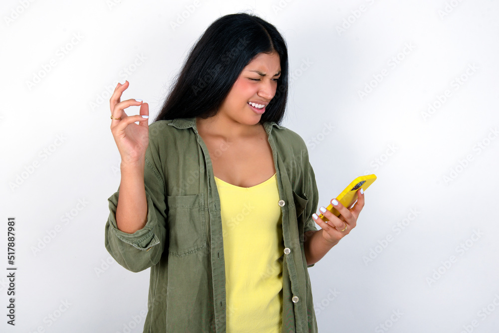 Photo of outraged annoyed young brunette woman wearing green overshirt standing against white wall holds cell phone, makes call, argues with colleague, expresses negative emotions. People and anger. Photo of outraged annoyed young brunette woman wearing green overshirt standing against white wall holds cell phone, makes call, argues with colleague, expresses negative emotions. People and anger.