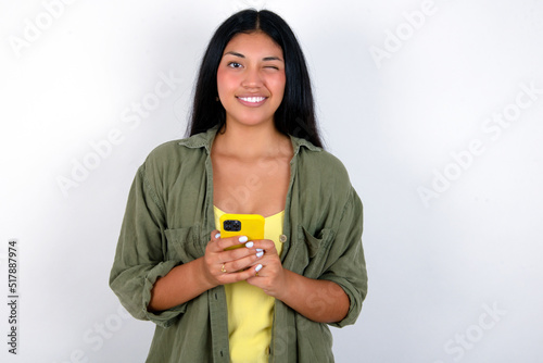 Pleased young brunette woman wearing green overshirt standing against white background using self phone and looking and winking at the camera. Flirt and coquettish concept.