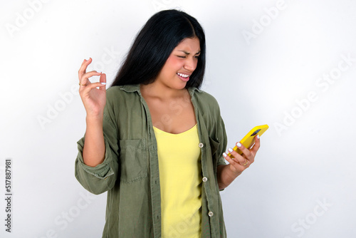 Photo of outraged annoyed young brunette woman wearing green overshirt standing against white wall holds cell phone, makes call, argues with colleague, expresses negative emotions. People and anger.
