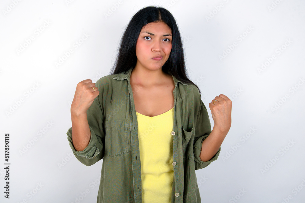 Irritated young brunette woman wearing green overshirt standing against white background blows cheeks with anger and raises clenched fists expresses rage and aggressive emotions. Furious model Irritated young brunette woman wearing green overshirt standing against white background blows cheeks with anger and raises clenched fists expresses rage and aggressive emotions. Furious model