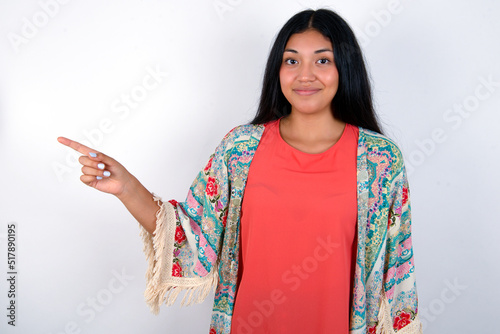 Positive young brunette woman wearing colourful outfit standing against white background with satisfied expression indicates at upper right corner shows good offer suggests to click on link