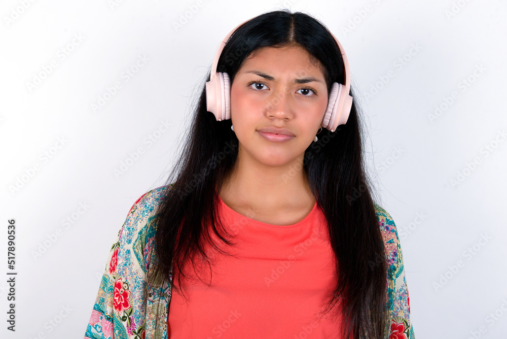 Serious displeased young brunette woman wearing colourful outfit standing against white background looks puzzled at camera being angry wears stereo headphones listens music while walking at street Serious displeased young brunette woman wearing colourful outfit standing against white background looks puzzled at camera being angry wears stereo headphones listens music while walking at street