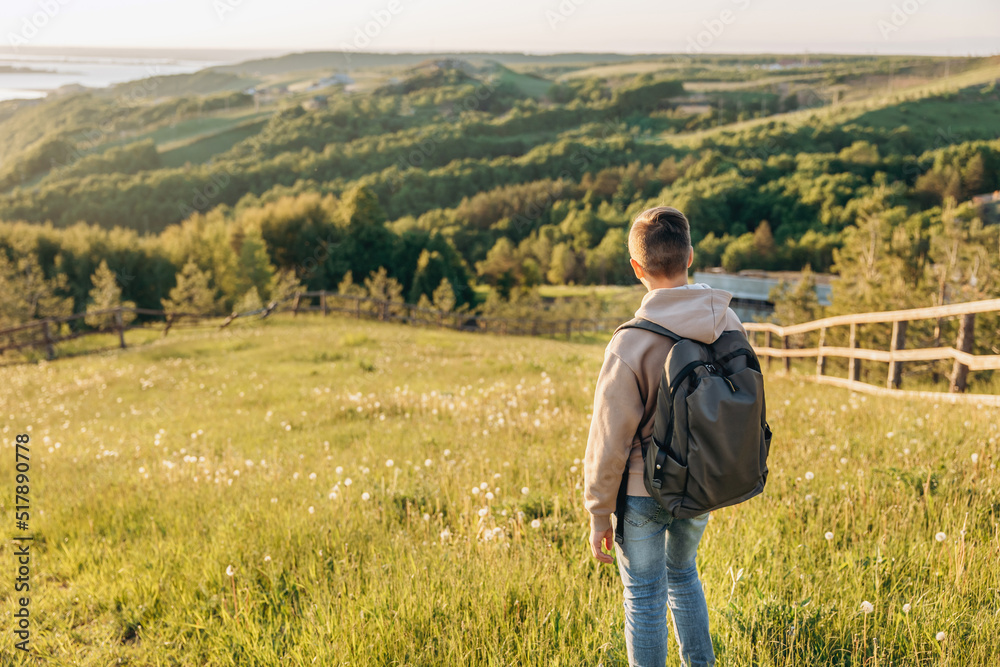 Tourist with backpack standing on top of hill in grass field and enjoying beautiful landscape view. Rear view of teenage boy hiker resting in nature. Active lifestyle. Concept of local travel Tourist with backpack standing on top of hill in grass field and enjoying beautiful landscape view. Rear view of teenage boy hiker resting in nature. Active lifestyle. Concept of local travel
