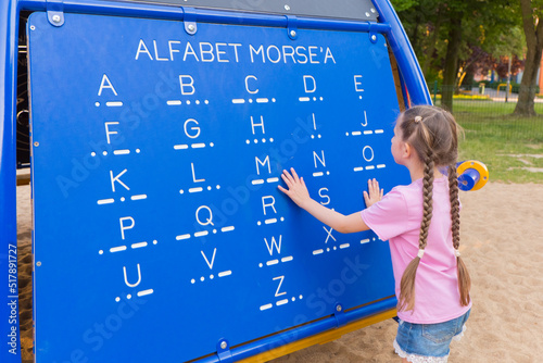 Photography A little girl 7-8 years old with long hair dressed in a pink T-shirt and shorts is studying Morse code on a blackboard in the playground