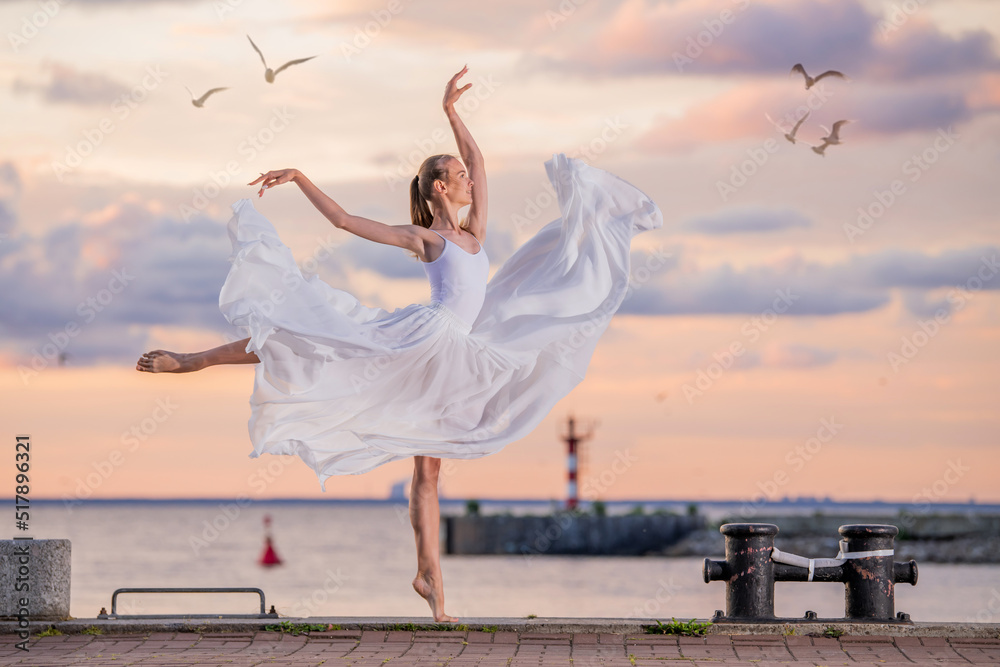 A dancing ballerina in a white flying skirt and tights on the ocean embankment or on the sea beach against backdrop of sunset sky A dancing ballerina in a white flying skirt and tights on the ocean embankment or on the sea beach against backdrop of sunset sky