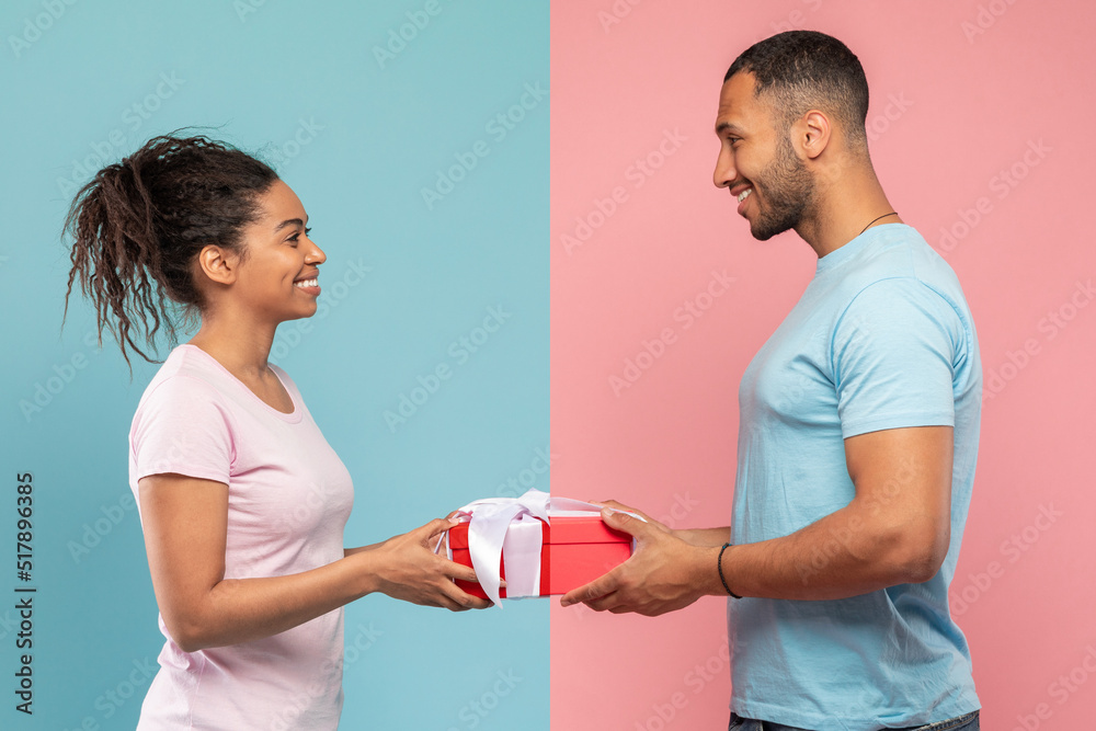 Loving black man giving gift to his wife, posing over blue and pink studio background, side view