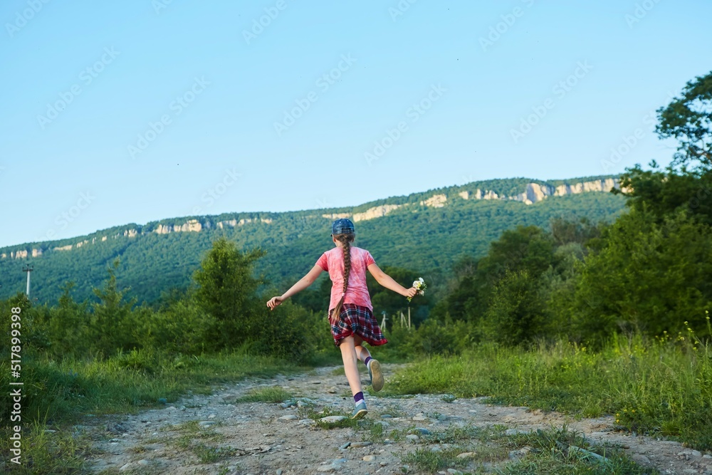Fototapeta premium A school-age girl in pink clothes runs along a dirt road and jumps for joy.