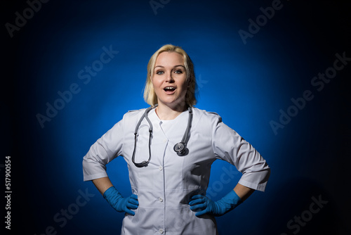 a female doctor in a white coat, gloves, with a stethoscope stands with her hands on her hips on a dark background