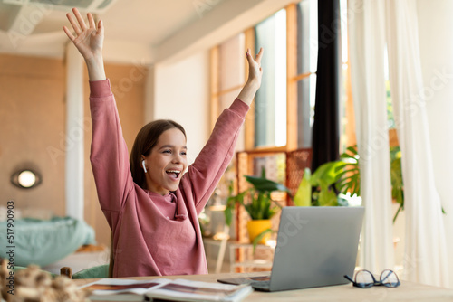 Emotional schoolgirl celebrating success, cheerful teenager raising hands up while using laptop at home, free space