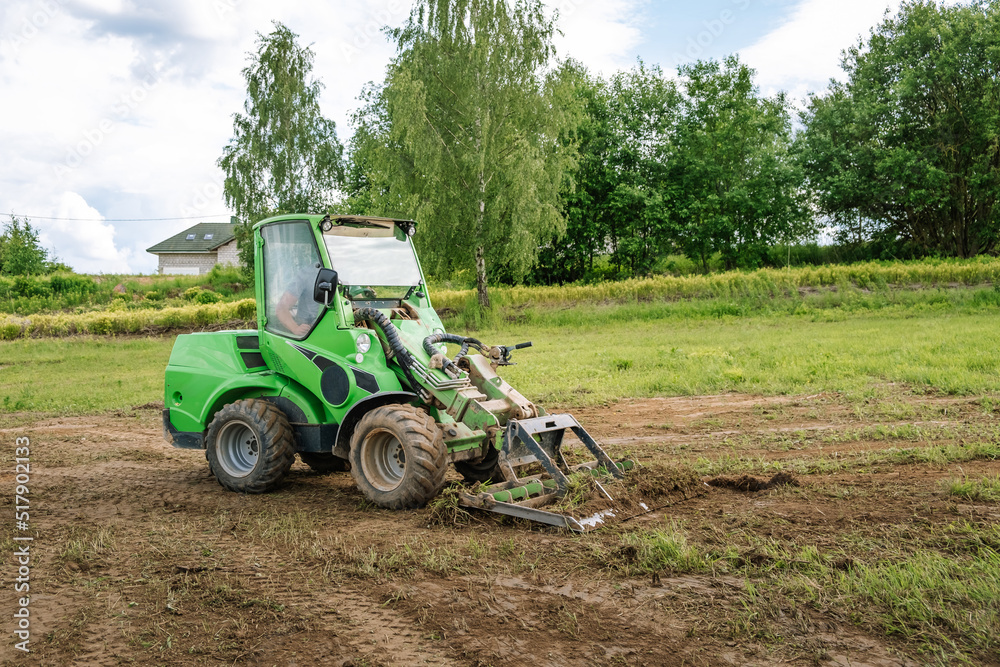 A green mini skid steer loader clear the construction site. Land work by the territory improvement. Small tractor with a ground leveler for moving soil, turf. Machine for agriculture work. Copy space A green mini skid steer loader clear the construction site. Land work by the territory improvement. Small tractor with a ground leveler for moving soil, turf. Machine for agriculture work. Copy space