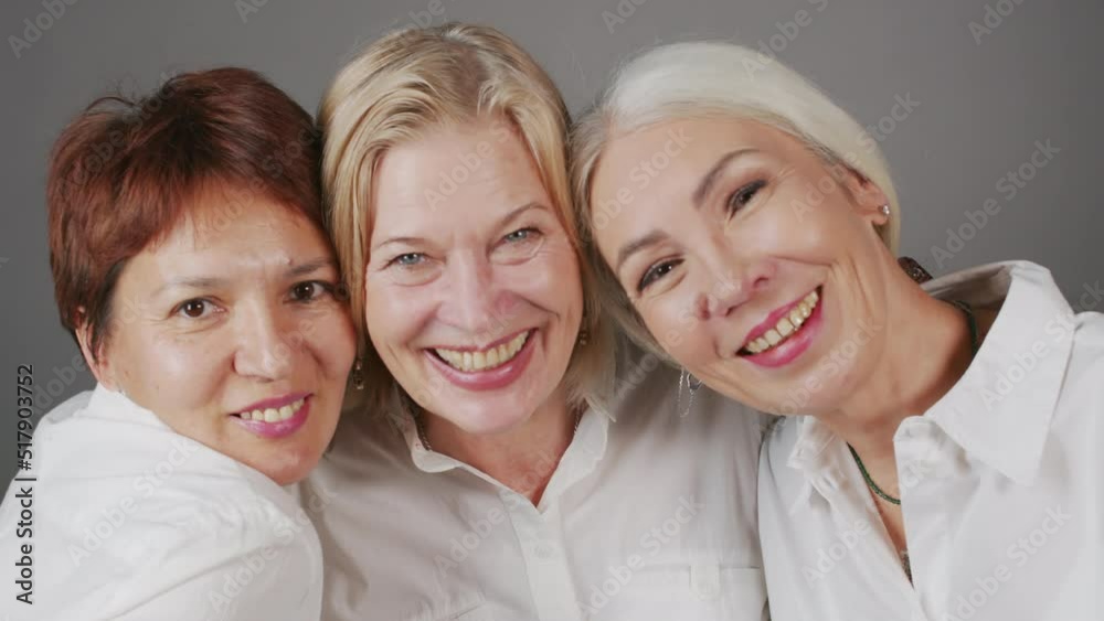 Chest up studio portrait of three cheerful mature girlfriends smiling at camera standing together on grey background