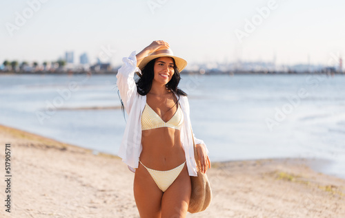 people, summer holidays and leisure concept - happy young woman in bikini swimsuit, white shirt and straw hat with bag walking along beach © Syda Productions