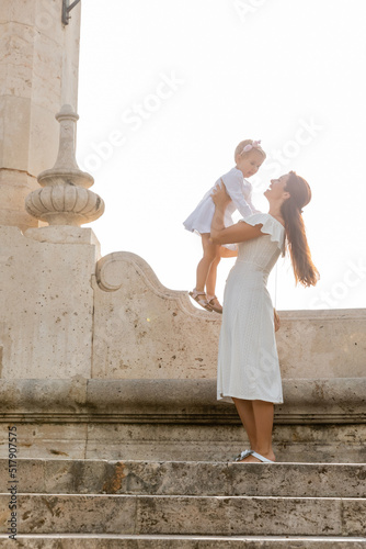 Smiling mother in dress holding daughter on stone stairs of Puente Del Mar bridge in Valencia.