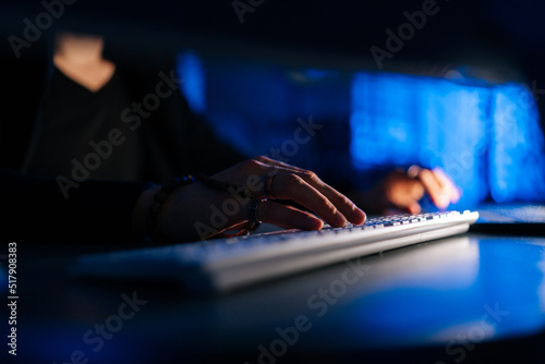 Close-up hands of unrecognizable hacker man working typing on keyboard laptop computer sitting at desk in dark room with blue neon lights. Concept of cyber attack, virus, malware, illegally.