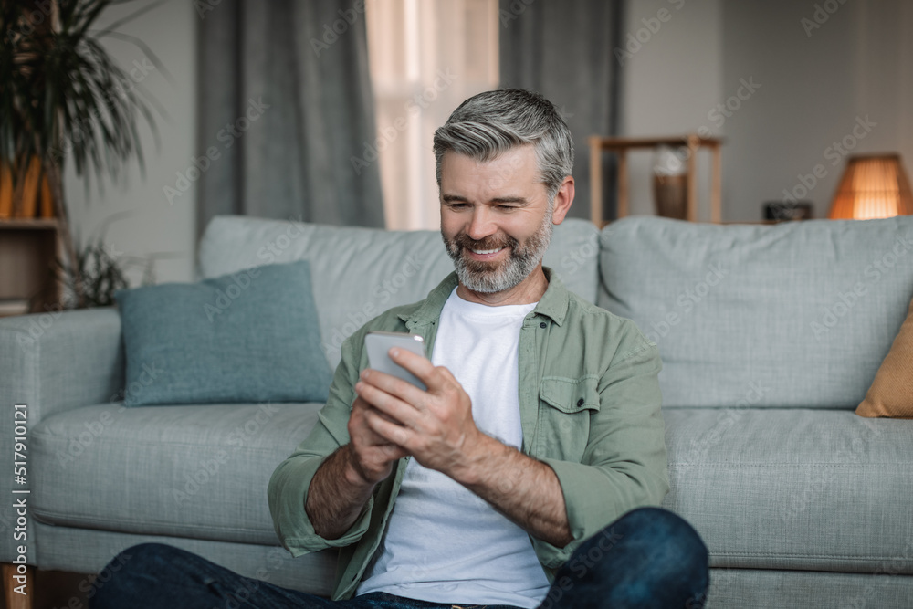 Happy elderly european man with beard typing on smartphone has chat in social media in living room