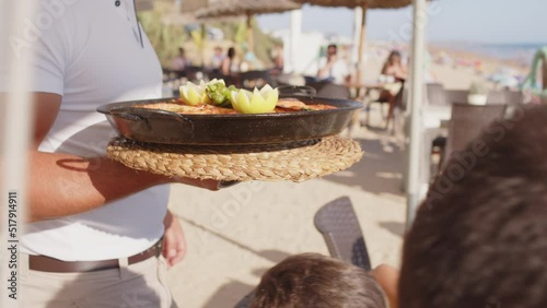 4K SlowMo - professional waiter bringing a plate of paella to a family's table in a restaurant on the beach