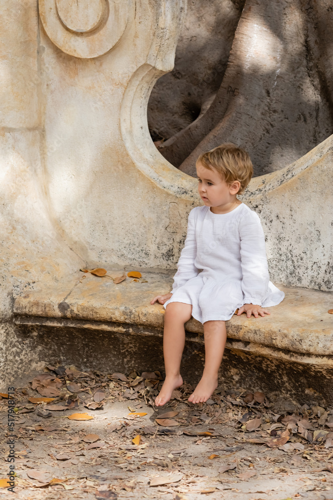 Barefoot baby girl in dress sitting on stone bench in park in Valencia. Barefoot baby girl in dress sitting on stone bench in park in Valencia.