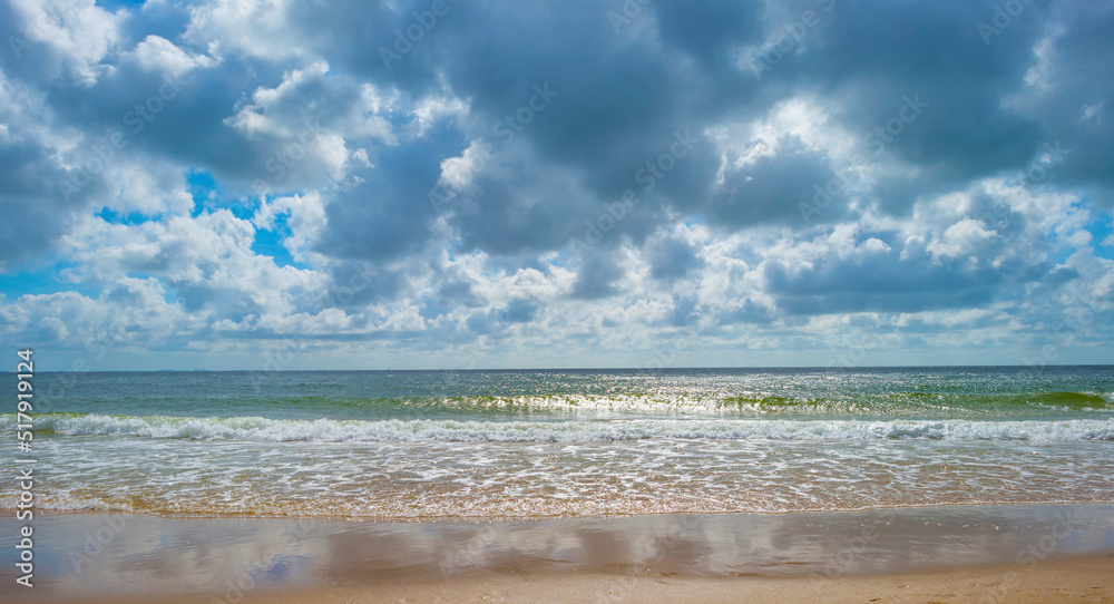 Sunlit waves and a wooden breakwater on the yellow sand of a sunny beach along a sea under a blue cloudy sky in summer, Walcheren, Zeeland, the Netherlands, July, 2022