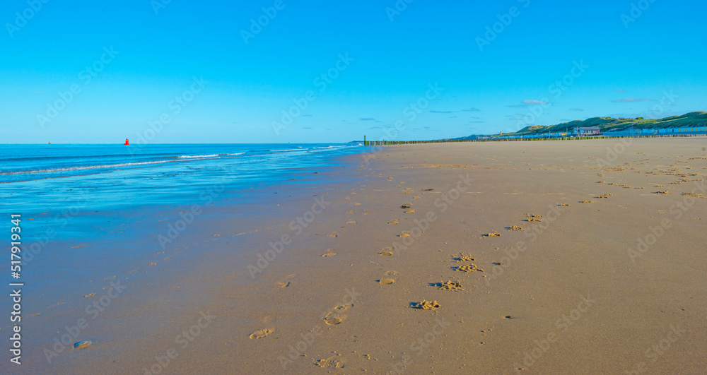 Sunlit waves and a wooden breakwater on the yellow sand of a sunny beach along a sea under a blue cloudy sky in summer, Walcheren, Zeeland, the Netherlands, July, 2022
