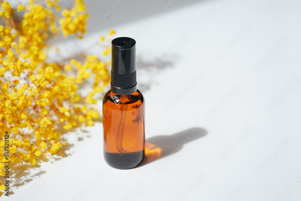 Amber pump bottle with serum, tonic or essential oil with yellow flowers. White background with daylight. Beauty concept for face and body care Amber pump bottle with serum, tonic or essential oil with yellow flowers. White background with daylight. Beauty concept for face and body care