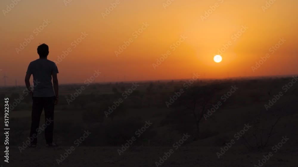 Rear view silhouette shot of an Indian man staring at the sun during the sunset. Man lost in deep thoughts looks at sunset. Man looks at the sunset and thinks of his future. New beginning background. Rear view silhouette shot of an Indian man staring at the sun during the sunset. Man lost in deep thoughts looks at sunset. Man looks at the sunset and thinks of his future. New beginning background.