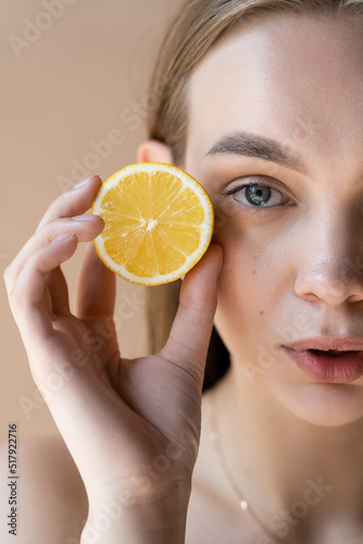 cropped view of woman with natural makeup holding half lemon isolated on beige.