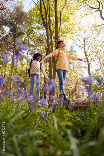 Low Angle Shot Of Two Children Walking Through Bluebell Woods In Springtime Balancing On Log