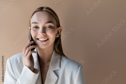 trendy woman smiling at camera while talking on mobile phone isolated on grey.