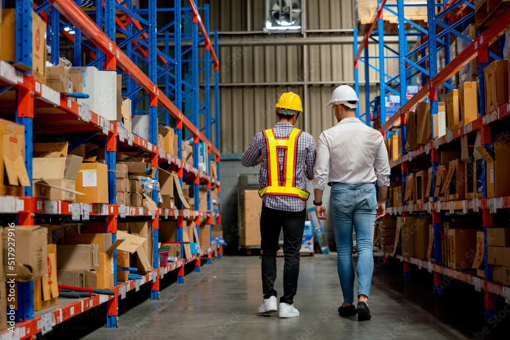 Back of two Asian and Caucasian warehouse workers walk and discuss on the way between shelves in workplace.