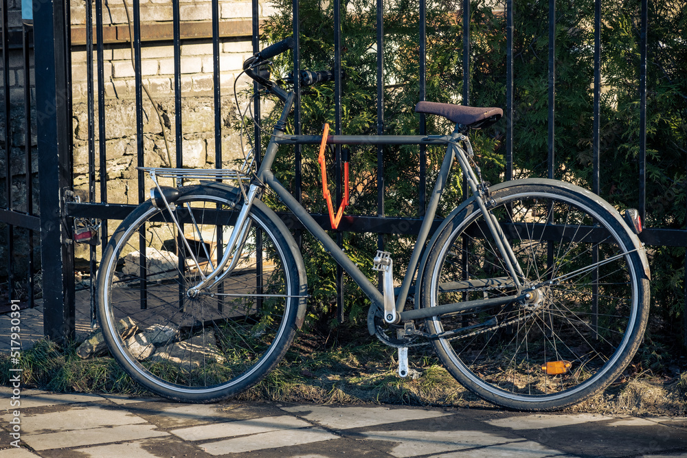 Black retro bicycle parked against metal fence with house wall and garden in the background. Summer, ecological, popular transport. Vintage bicycle with lock. Safe parking in the city