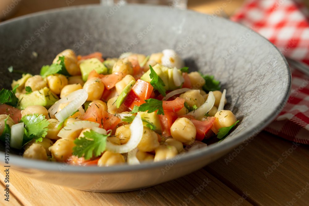 45º view of a chickpeas salad with avocado, tomato, coriander and onion, served on a grey dish with a red chekered cloth on a wooden table. Horizontal image 45º view of a chickpeas salad with avocado, tomato, coriander and onion, served on a grey dish with a red chekered cloth on a wooden table. Horizontal image