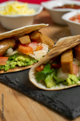 Closeup of two vegan guacamole and seitan tacos served on a blackboard with lime and pico de gallo, on a wooden table with a red cloth behind. Vertical image