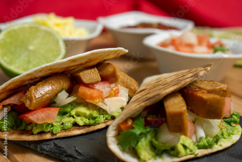 Side view of two vegan guacamole and seitan tacos served on a blackboard with lime, cheese and pico de gallo, on a wooden table with a red cloth behind. Horizontal image