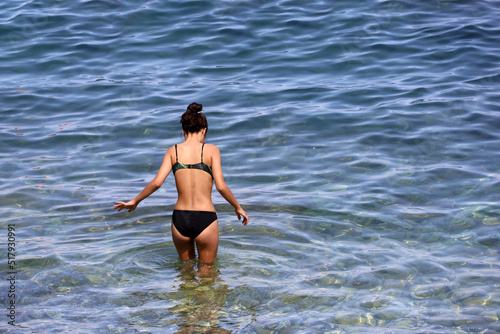 Slim girl in black swimsuit going by rocky bottom to swim in blue transparent water. Vacation on summer beach