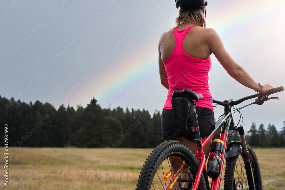 A woman cyclist stands next to her mountain bike and admires the rainbow in the background. A woman cyclist stands next to her mountain bike and admires the rainbow in the background.