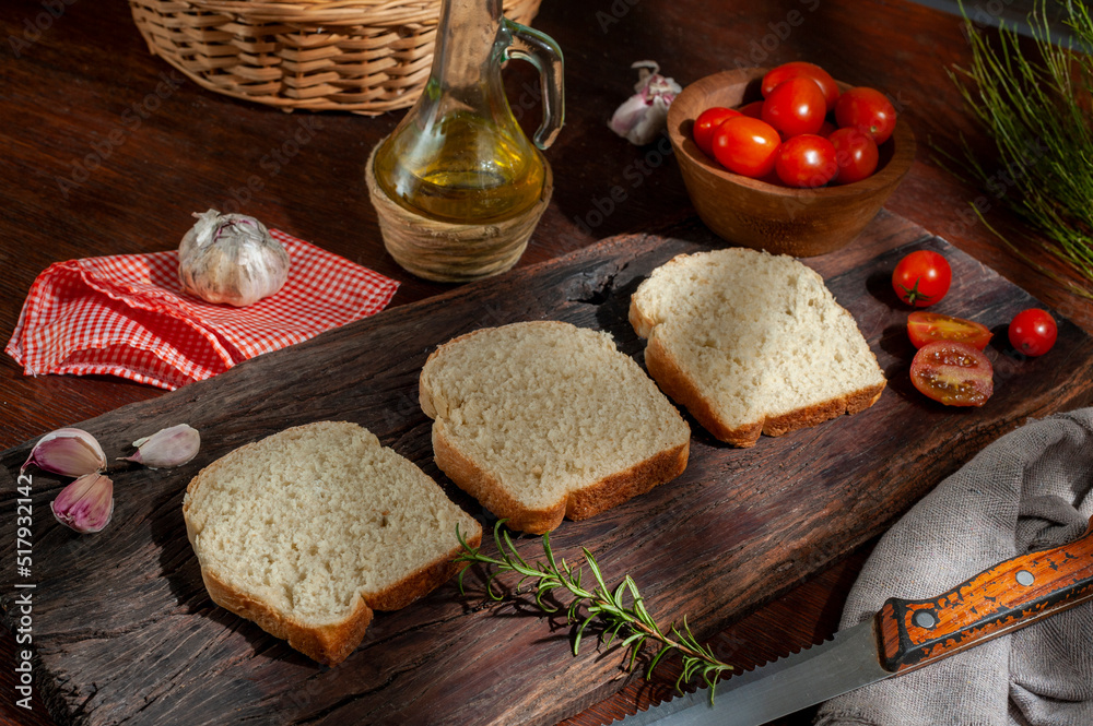fresh and homemade whole and sliced bread with oil can and cherry tomatoes wicker pan and rosemary on wooden table with light from a window